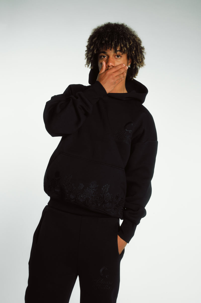 Model wearing the Hoodie Shadow Tracksuit - Bloom Hoodie in black with floral embroidery, showcasing relaxed fit and cotton texture.