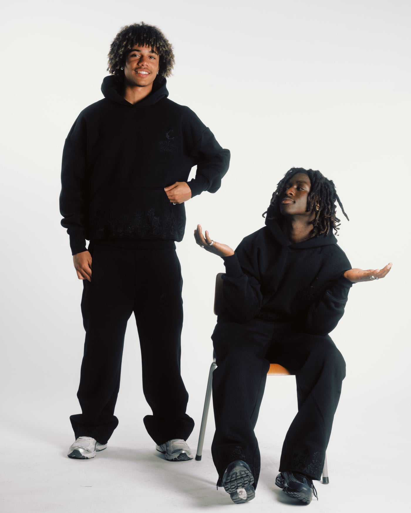 Male models in Hoodie Shadow Tracksuit - Bloom Hoodie set, styled in black cotton with floral embroidery for modern streetwear appeal.